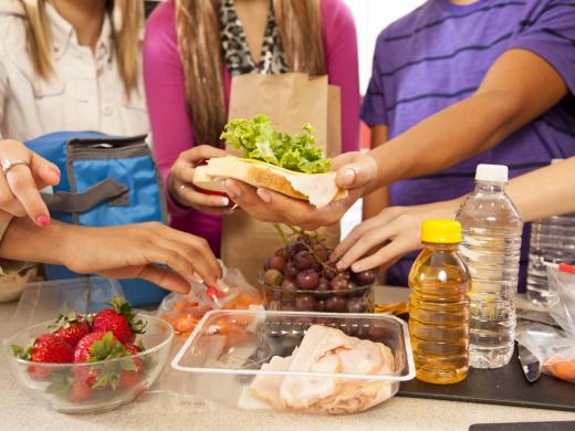 A group of young people gather around a counter where they are preparing healthy packed lunch together, including a turkey sandwich, baby carrots, strawberries, and graps.