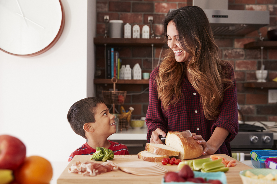 A mother smiles as she slices a loaf of bread on a counter and looks down at her son who is smiling back at her.