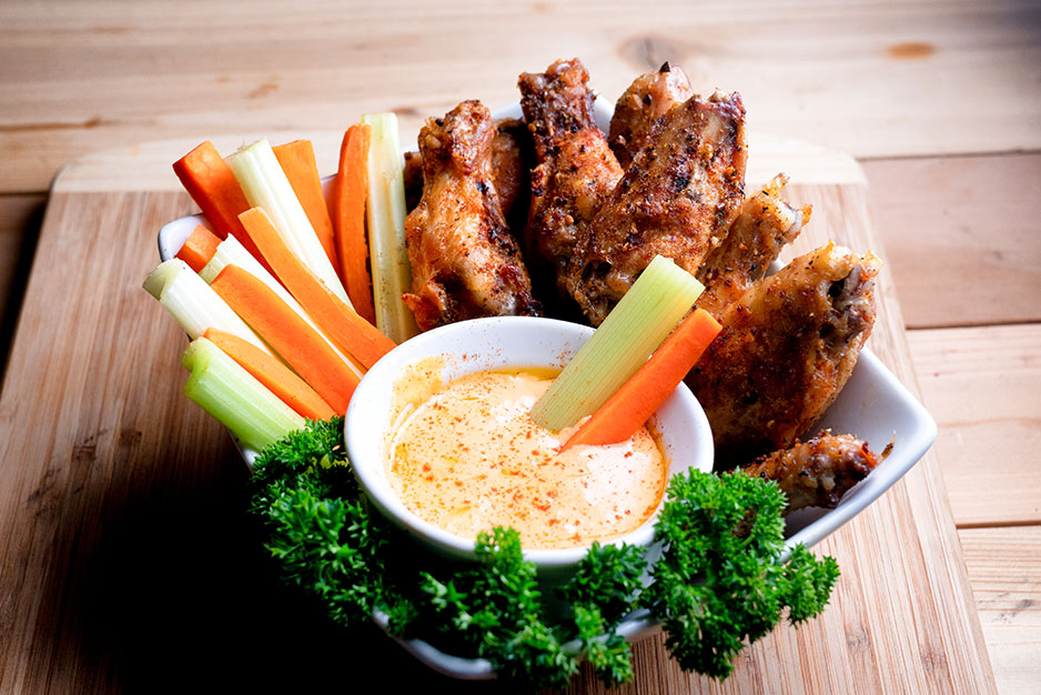 A dish with celery, carrot sticks, and chicken wings and  a bowl of buffalo sauce.
