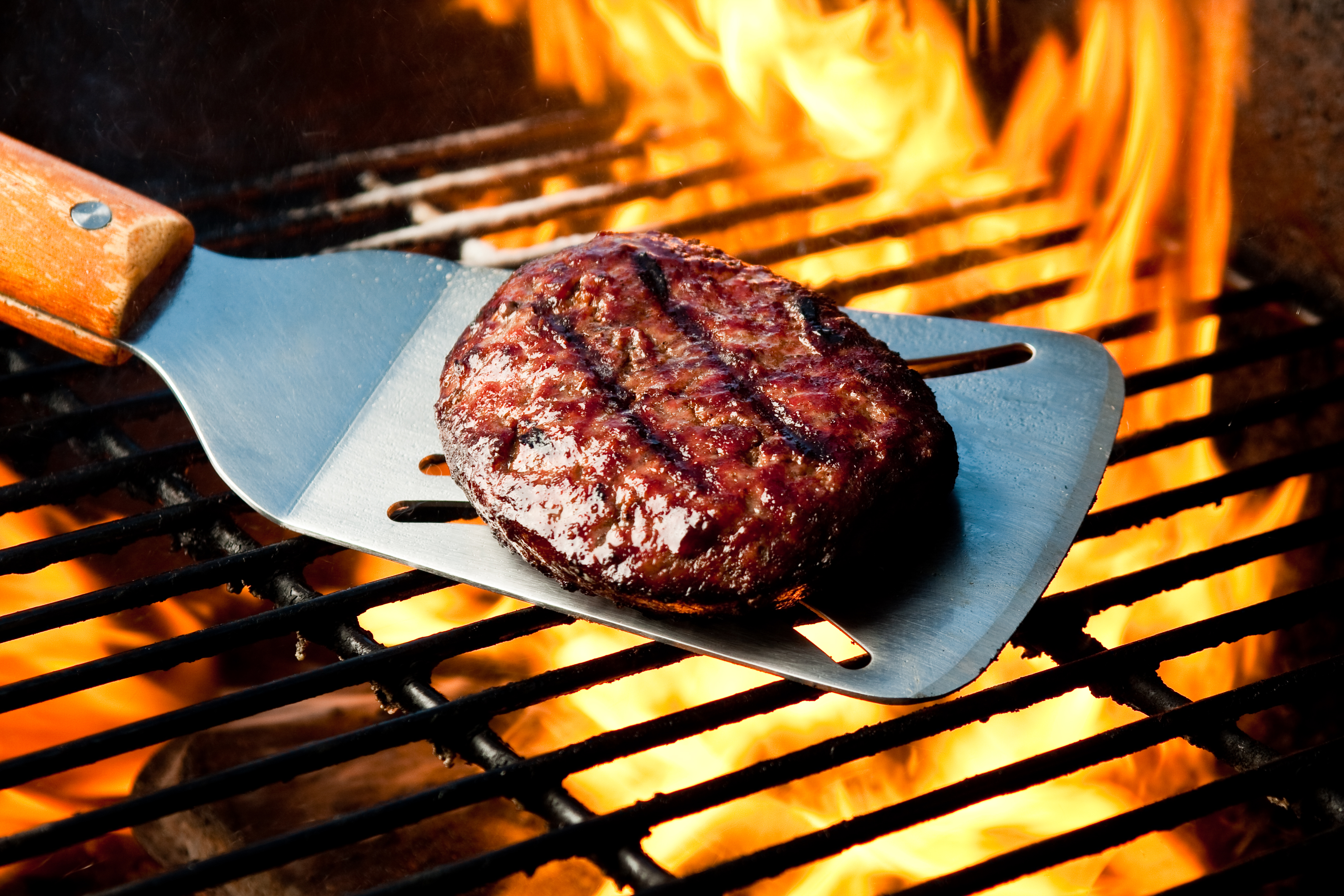 A seared hamburger on a grill spatula over a flame.