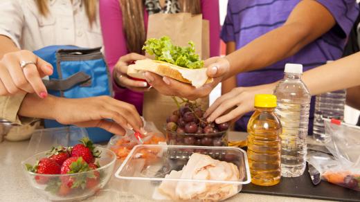 A group of young people gather around a counter where they are preparing healthy packed lunch together, including a turkey sandwich, baby carrots, strawberries, and graps.