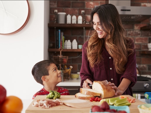 A mother smiles as she slices a loaf of bread on a counter and looks down at her son who is smiling back at her.