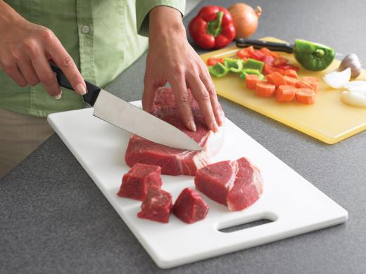 Una persona cortando carne cruda sobre una tabla de cortar. En otra tabla de cortar se ven zanahorias, pimientos y cebollas picados.