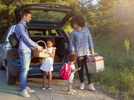 A family with two young children unpack coolers and snacks from the back of their car.