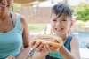 A Mom and a boy at a pool. The smiling boy is being handed a hot dog.